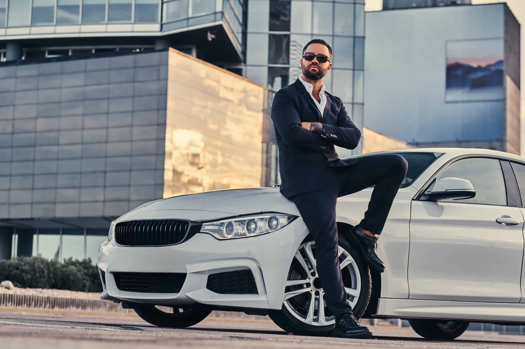Bearded male in sunglasses dressed in a black suit sitting on luxury car against a skyscraper.